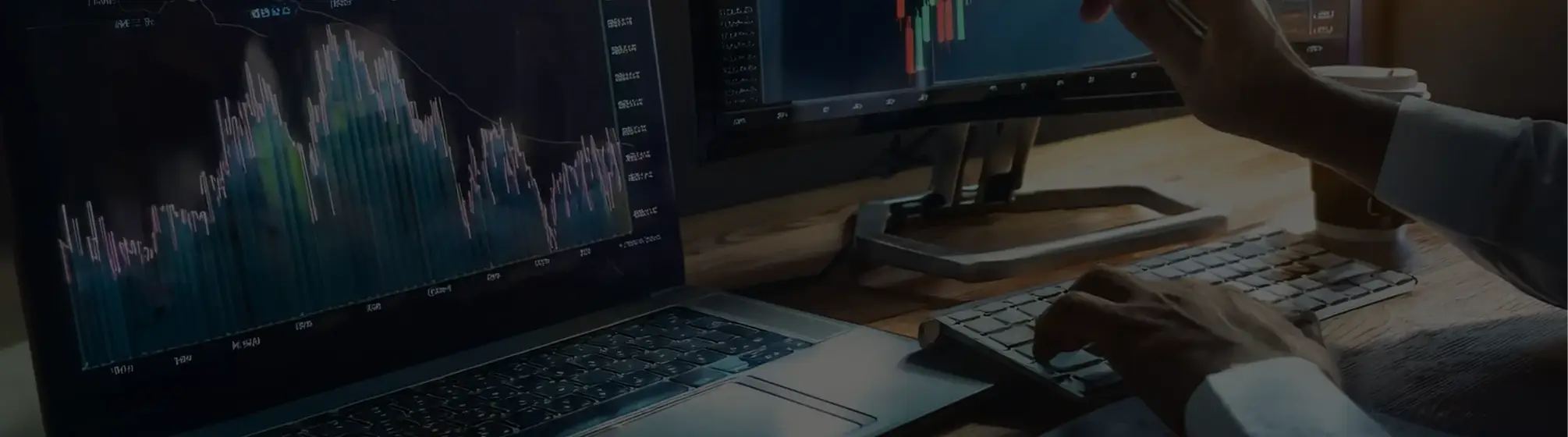 A close-up of a trading desk with dual monitors displaying colorful financial charts, next to a laptop and a wireless keyboard.