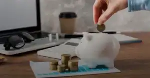 Hand deposits euro coin into white piggy bank on desk with laptop, coins, calculator, and chart; symbolizes saving, finance, and budgeting.
